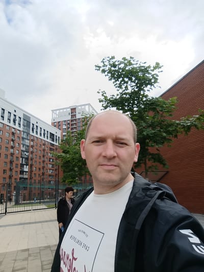 The image is a selfie taken outdoors in Naro-Fominsk, Russia. The foreground is dominated by a man in a black hooded jacket over a white t-shirt. He is looking towards the camera with a neutral expression. The t-shirt has text and graphics printed on it, but the text is partially obscured and reversed due to the selfie perspective. Some of the visible text on the t-shirt appears to be in a European language, possibly English.
In the background, there are several modern residential buildings with a brick facade and white accents. One building on the left is taller and has numerous windows. Another building on the right is shorter and appears to be a contrasting architectural style. Green trees are interspersed between the buildings, and a dark metal fence is visible in front of the buildings. A woman in a dark jacket and patterned shirt is standing behind the fence, looking away from the camera. The sky is overcast with clouds, suggesting a cloudy day. The paving stones on the ground indicate an urban or suburban environment, possibly a public walkway or courtyard. There are no clear indicators of the time of day, but the lighting suggests it is daytime. The overall scene is one of a typical urban residential area.