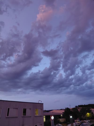 The image shows a view of Ecublens, Switzerland, during dusk. The sky is filled with dramatic purple and pink clouds, with a hint of lighter blue peeking through. In the foreground, a modern, light-colored, multi-story building with several rectangular windows is visible on the left. One window on the right side of this building is illuminated with a warm yellow light. In the background, a treeline and other buildings are faintly visible. A street with several parked vehicles, including vans and cars, is also present in the lower right portion of the image. A sign with a prominent 'e' logo is visible on the left side of the background, potentially indicating a commercial area.