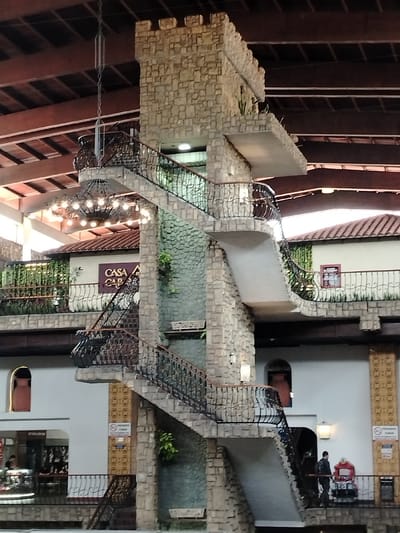 A stone tower dominates the image, incorporating a series of staircases with wrought-iron railings. The tower seems to be part of an indoor structure reminiscent of a courtyard. Buildings with balconies and tiled roofs flank the central tower. A chandelier hangs above, and people can be seen near the base of the tower. The overall scene has an architectural design blending castle-like elements with an indoor shopping or public space.