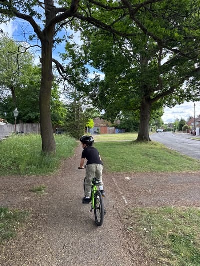 A boy wearing a black helmet, a black shirt, and camouflage pants is riding a green bicycle on a path. The path is lined with grass and large trees, with a street visible in the background. The location is Hazlemere, United Kingdom.