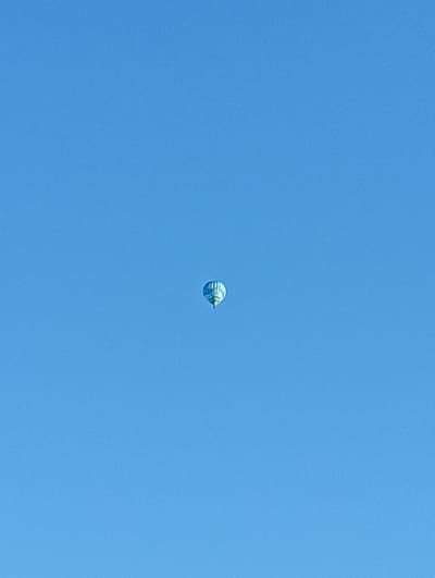 A hot air balloon, patterned with blue and white, is visible in the clear blue sky over Zofingen, Switzerland. The balloon is in the center of the frame and appears to be at a considerable altitude, with no other objects or people visible in the image. The sky is uniformly blue, indicating a clear and sunny day.