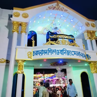 The image captures a brightly lit, ornate entrance gate to a fair or festival at night. The gate is predominantly white with extensive gold decorative elements, including intricate carvings on columns, archways, and border trim. Two large, multi-tiered crystal chandeliers hang, one prominently in the upper section and another below the main archway, adding to the festive atmosphere.

The structure features an upper tier with arched openings, illuminated by colorful LED lights (blue, red, green, white) arranged in a grid pattern. Similar colored lights are embedded around the main archway at ground level.

Prominently displayed on the main archway in blue Bengali script is the text: "চরফ্যাসন শিল্প ও বাণিজ্য মেলা-২০২৩", which translates to "Charfasson Industrial and Trade Fair - 2023". This indicates the event and year.

A crowd of people is visible at the ground level, passing through the entrance. They appear to be casually dressed attendees of the fair. Beyond the gate, the background reveals more vibrant, colorful lights and structures, suggesting the lively ambiance of the fair stretching into the distance. The dark sky above the illuminated gate confirms it is nighttime.