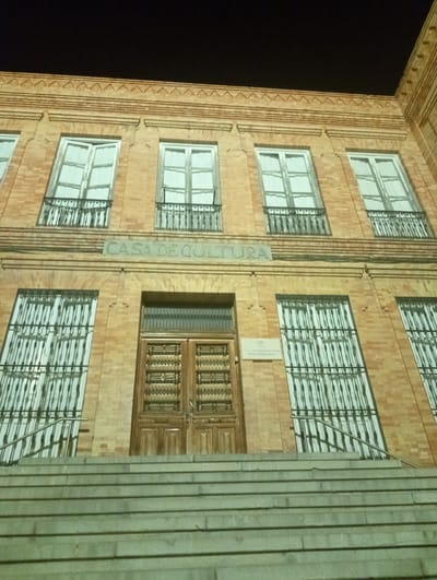 The image shows a low-angle shot of the facade of a brick building at night. The building has multiple stories with large, white-curtained windows, many of which are protected by wrought-iron grates. A prominent sign above the central double doors reads "CASA DE CULTURA," indicating it is a cultural center. A set of wide stone stairs with metal railings leads up to the entrance. The sky above is dark, suggesting it is nighttime. The building's architecture features decorative brickwork along the roofline. There are no people visible in the image. This building is the Casa de Cultura in Priego de Córdoba, Spain.