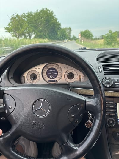 The image shows the interior of a Mercedes-Benz car, likely a W211 E-Class, from the driver's perspective. The steering wheel, dashboard, and instrument cluster are clearly visible. The instrument cluster displays information such as speed (30 km/h), engine temperature (+27.5°C), and odometer reading (432830 km). There is a key fob with a Mercedes-Benz logo hanging from the steering wheel. The car appears to be driving on a road with trees and vegetation on either side. A speed limit sign (60) is visible on the right side of the road. The sky is overcast, and there are raindrops on the windshield. Based on the Google Street View imagery of Yoakim Gruevo, Bulgaria, the road visible in the image is likely to be within the vicinity of the town, possibly on the road leading towards or away from it. The landscape suggests a rural or semi-rural setting. No people are visible in the image, other than the implied presence of the driver.