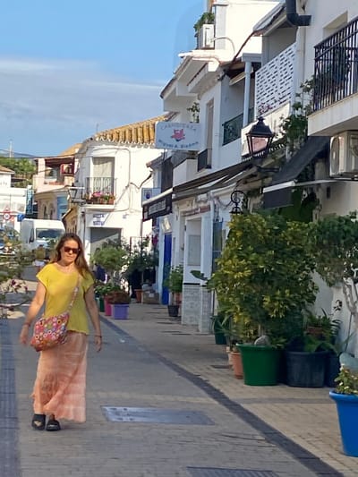In Cancelada, Spain, a woman walks down a sunlit pedestrian street lined with white buildings. She is wearing a yellow t-shirt, a long, flowing peach-colored skirt, and dark sandals. A colorful patterned bag is slung across her shoulder. Her hair is long and brown, and she wears sunglasses. In the background, shops with awnings are visible, including one with a sign that reads "CARNICERIA Miguel A. Biedma". Various potted plants are placed along the street, some containing vibrant flowers. A white van is parked further down the street. The sky is blue with scattered clouds.