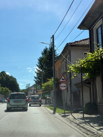 The image depicts a view from inside a vehicle traveling down a street in Beli Osam, Bulgaria, on a bright, sunny day. The road is paved and appears to be a two-lane street. In the foreground, the rear of a light-colored station wagon is visible, with license plate "OB 9023 BH". Ahead of it, another similar car with license plate "OB 0776 BB" is also on the road.
The street is lined with buildings on the right side, characteristic of a European village. One building, in the middle ground, is a light-colored, two-story structure with a tiled roof. A vine with green leaves is growing on the side of this building, partially obscuring it. There are several street signs on a pole near this building. The uppermost sign is a red and white triangular sign with a black wavy line, indicating a winding road. Below it is a circular red and white sign with two black car silhouettes, signaling no overtaking.
On the left side of the street, lush green trees and vegetation are visible, with mountains in the background. Power lines are strung across the sky, supported by utility poles. The sky is clear blue with a few scattered white clouds. The overall impression is of a quiet, rural setting within the village of Beli Osam.