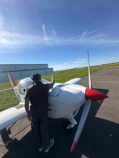 The image depicts a person performing what appears to be a pre-flight inspection or preparation on a small, white, single-engine propeller aircraft at an airfield. The person, seen from behind, is wearing a dark vest over a long-sleeved shirt, dark trousers, and sneakers, with a cap on their head. Their right arm is extended towards the aircraft's cockpit area, suggesting engagement with the plane.

The aircraft is white with a distinctive red propeller spinner and red tips on its propeller blades. On the wing, the text "Aéroclub Air F[rance]" is clearly visible, identifying the plane's affiliation. The plane is parked on a dark asphalt tarmac, which is bordered by a vibrant green grassy area. In the distance, another small white aircraft can be seen on the grass, and some hangars or similar buildings line the horizon.

The scene is set under a clear, bright blue sky with a few wispy white clouds and prominent contrails, indicating good weather conditions. Strong, well-defined shadows are cast on the tarmac by the aircraft and the person, suggesting direct sunlight, likely during the morning or late afternoon. The overall atmosphere conveys a sense of calm and operational readiness at an airfield, specifically in Toussus-le-Noble, France.