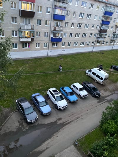 The image depicts a residential building in the city of Beloretsk, Russia. The building is a multi-story apartment complex with light-colored concrete facade. There are several windows and balconies visible. In front of the building, there is a grassy area and a paved parking lot. Several cars are parked in the lot, including a white van, a white SUV, a black sedan, a white hatchback, a blue sedan, and a dark-colored sedan. Two people are walking on the grass in the middle ground. One person is sitting on a bench near the white van. The ground appears damp, suggesting recent rain or puddles. The overall scene suggests a typical urban residential environment.