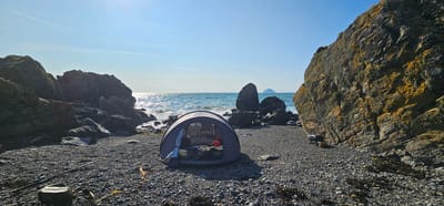 This is a photograph of a rocky beach in Lendalfoot, United Kingdom. The scene is bathed in bright sunlight. In the foreground, a gray pop-up tent is set up on the pebble beach. Inside the tent, sleeping bags and other camping gear are visible. To the right of the tent, a small portable grill sits near a large, lichen-covered rock formation. Fishing rods are laid out on the pebbles in the mid-ground. The ocean is visible behind the tent, with waves gently lapping against a cluster of large rocks in the water. In the distance, a small island or sea stack is visible on the horizon. The sky is clear blue with faint contrails from an airplane. The overall impression is one of a serene and isolated coastal camping spot.
