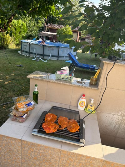 In a backyard setting that appears to be in Bozhurishte, Bulgaria, a barbecue is underway. Raw, marinated meat is arranged on a portable electric grill. Nearby, stacked plastic containers hold more uncooked meat. A green glass bottle of beer stands beside the grill. In the background, an inflatable swimming pool is set up, with two individuals enjoying a dip. Lush green trees and foliage surround the area, suggesting a sunny day. The scene conveys a relaxed, outdoor dining and recreation atmosphere.