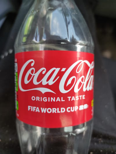 The image shows a close-up of a clear plastic Coca-Cola bottle with a red label. The label prominently displays the Coca-Cola logo in white script. Below the logo, the text "ORIGINAL TASTE" is visible in white capital letters, followed by "FIFA WORLD CUP 20" in the same font and color. The bottle appears to be partially filled with a clear liquid. The background is dark and out of focus, suggesting an indoor or dimly lit environment. There are no people or specific landmarks identifiable that would indicate a location in Madan, Bulgaria.