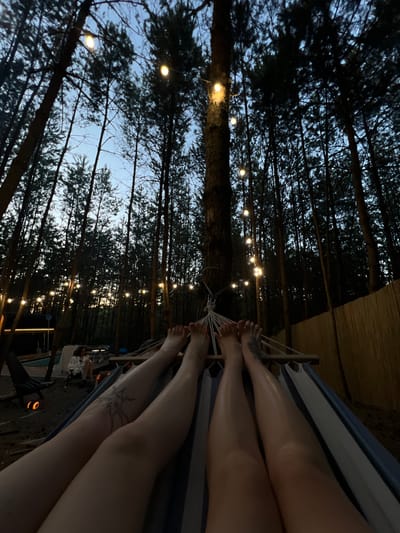 In Ivachiv Horishnii, Ukraine, two individuals' legs are seen resting in a hammock within a forested area at dusk. String lights are hung between tall trees, illuminating the twilight sky. In the background, a person sits near a fire pit, and other outdoor furniture is visible. The overall scene depicts a relaxed evening in a natural setting.