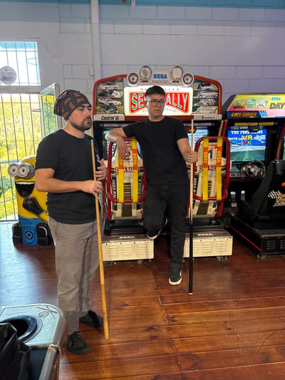 Two men hold pool cues horizontally in front of two racing arcade games with "SEGA" and "RALLY 2" on the marquees. The man on the left wears a bandana and a t-shirt with gray pants. The man on the right wears glasses and a t-shirt with black pants. A minion figurine sits next to the left arcade game. The room has wooden floors and blue-and-white walls. A window is on the left with outdoor vegetation visible. This is likely a recreation center or arcade game room. The text on the window reads "PROPIEDAD PROTEGIDA". The location appears to be Villa General Belgrano, Argentina based on the text on the bandanna of the person on the left: "ROYAL ENFIELD".