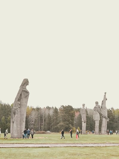 The image shows the Salaspils Memorial Ensemble in Mežezeri, Latvia. Five large, grey stone sculptures stand in a field of grass. People are walking around the sculptures and laying flowers at their bases. A forest is in the background. The sky is overcast.