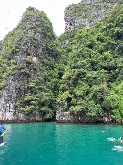 The image depicts a scenic view of a limestone cliff covered in lush green vegetation, bordering a vibrant turquoise body of water. Several people are swimming and enjoying the water. The sunlight suggests it is daytime. The scene evokes a sense of relaxation and recreation. The location is identified as Haad Yao, Thailand.