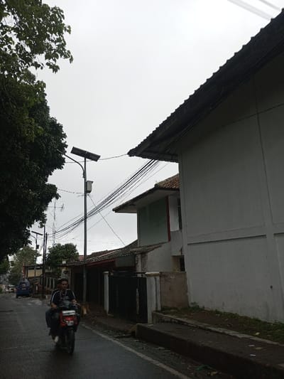 The image depicts a street scene in Ambakirano, Madagascar, under an overcast sky. A lone male individual is operating a red two-wheeled scooter on a wet asphalt road, traveling towards the viewer's left. He wears a dark shirt. To the right, a multi-story building with a light grey facade and a tiled roof is prominently featured. A white piece of cloth hangs from an upper section of this building, near a lighter green-painted wall. Utility poles equipped with solar panels and luminaires line the left side of the street, carrying numerous black electrical cables that stretch across the scene, connecting to buildings and other poles. Dense green foliage from trees partially obscures the upper left portion of the sky. Further down the street, additional residential-style buildings with tiled roofs and various fences are visible, along with a blue vehicle parked on the left side of the road. The road surface appears wet, indicating recent precipitation.