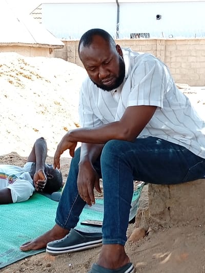The scene is set outdoors in Gboko, Nigeria, depicting two males. In the foreground, a dark-skinned male with short dark hair and a beard sits on a low, rough earthen block. He wears a short-sleeved white shirt with subtle vertical stripes and dark blue jeans. He is barefoot, with a dark green slip-on shoe featuring white stripes resting on the ground near his right foot, and a similar shoe partially visible on his left foot. His head is bowed, and he displays a solemn expression. To his left, another dark-skinned male lies supine on a light green woven mat on the ground. He wears a white short-sleeved shirt, with his left arm bent and hand near his head, looking upwards. The ground is composed of loose, light brown earth. In the mid-ground, a substantial pile of light brown sand or earth is visible. The background features a light-colored building with a corrugated metal roof on the left, and a light-colored concrete block wall topped with barbed wire on the right.
