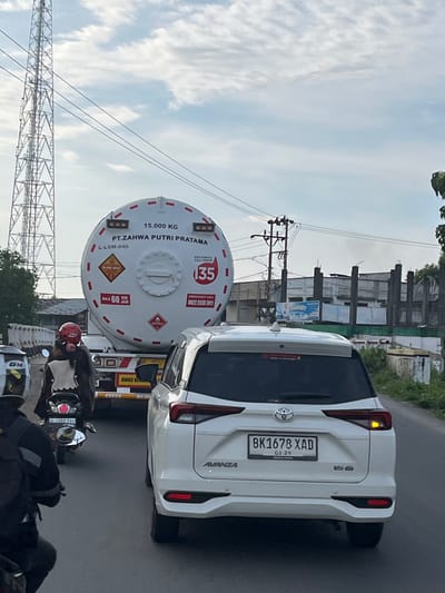 The image captures a street scene in Uleemadon, Indonesia, characterized by moderate traffic and a clear sky with scattered clouds. In the foreground, a white SUV with the license plate BK1678 XAD is prominently featured, its brake lights illuminated. To the left of the SUV, a motorcycle with a rider wearing a red helmet is visible, moving in the same direction. Behind the SUV, a large white tanker truck with "PT.ZAHWA PUTRI PRATAMA" written on it is partially obscured. The tanker truck has markings indicating a capacity of "15.000 KG" and a speed limit of "MAX 60 KM JAM." Further back, a tall communication tower stands against the sky, and various buildings with unfinished construction are present in the background. Power lines are visible throughout the scene, indicating the urban or semi-urban environment.