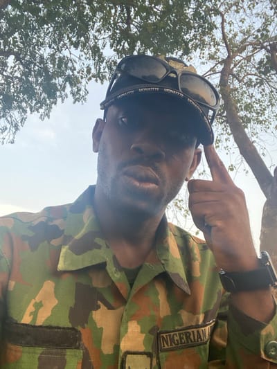 A low-angle shot features a person in a Nigerian military camouflage uniform. "NIGERIAN" is stitched above a Velcro patch on the right breast. Sunglasses, branded "MONATTE," are perched on their forehead. The person points towards their temple with their right index finger. A black smartwatch is visible on their wrist. Trees and a bright sky are in the background.