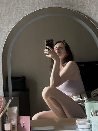 A young woman is taking a selfie in a mirror, reflecting her upper body and legs as she sits with her knees drawn up. She is wearing a light purple camisole and grey shorts. She holds a black smartphone in her right hand, with her left hand resting on her chin. The mirror is large and arched, framed by a light-colored border, and positioned against a textured grey wall. In the foreground, on a surface below the mirror, various cosmetic items are visible, including a pink notepad with Korean writing, a tube of cream, and a small white jar. The lighting is dim, suggesting an indoor setting, likely a bedroom or dressing area. The reflection shows a portion of a bed and a dark object that could be a pillow in the background. There are no indications of the city of Dongducheon-si, South Korea.