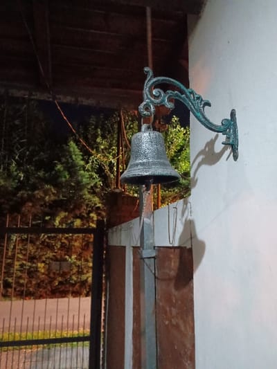 A metal doorbell with an ornate, decorative, blue-green finish is mounted on the exterior wall of a building. It is positioned to ring a bell. The bell is a circular, metal object with a dark, silver-grey finish. It is suspended from the mounting bracket and is angled downwards. The wall is painted white. The building is located in El Talar, Argentina. The latitude and longitude coordinates are -34.473 and -58.639. The image appears to be taken at night. The background is dark and blurred. The doorbell is the main subject of the image. There are no people visible. There is a large, metal gate in the background. The gate is open. There are trees and foliage visible in the distance. The lighting is dim. The image is well-composed and sharp. The image is taken from a low angle. The image is taken from a distance.