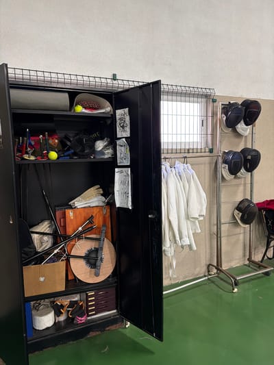 The image depicts an indoor storage area within a sports facility, specifically a fencing club, located in Santarém, Portugal. A large, black metal storage cabinet, with its right door ajar, occupies the left side of the frame. Inside the cabinet, multiple shelves hold various items: the top shelf contains a grey mat, a yellow tennis ball, a conical object filled with small red items, and a white cap; the second shelf includes several tools, another yellow tennis ball, a black fan-like object, and items in clear plastic bags; the third shelf features cream-colored protective gloves, a brown wooden case, and multiple long, slender poles or swords; the bottom shelf holds a cardboard box, a large wooden disc with a black handle, white cylindrical weights, a blue object, and a multi-tiered brown drawer unit. Taped to the inside of the cabinet door are several pieces of paper with abstract black markings.
To the right of the cabinet, a metal garment rack displays multiple white fencing jackets hanging on individual hangers. Further to the right, a vertical metal rack holds six fencing masks, primarily black with white interior padding, arranged in three pairs. A red bag is partially visible behind the mask rack. The background consists of a light-colored, textured wall, with a barred window visible above the hanging jackets. A green, smooth floor covers the foreground. A metal mesh fence runs along the upper part of the wall, extending across the top of the cabinet and the window. No individuals are present.
