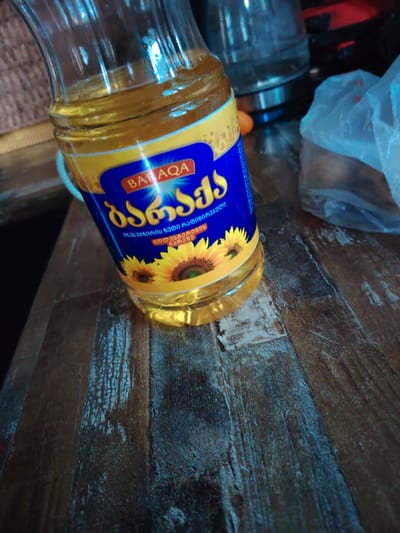 A bottle of sunflower oil with a yellow and blue label featuring sunflowers is placed on a wooden table. The label has Georgian script and the brand name "BARAQA". The bottle is partially filled with golden oil. The table surface is dark and has visible wood grain and some distressed textures. In the background, out of focus, are other objects including what appears to be a kettle or similar appliance and a plastic bag. The lighting suggests an indoor setting, possibly in a kitchen or dining area. The image is taken from a slightly high angle, looking down at the bottle and table. There are no people or animals visible in the image. The scene is static and depicts a simple still life arrangement. The location is not explicitly stated in the image, but the script on the label is Georgian.