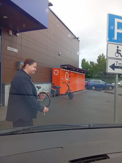 A woman with brown hair tied back walks away from a red delivery locker facility. She is wearing a dark gray jacket and is holding onto the handlebars of a bicycle that is being pushed along. The locker facility has the brand name "Omniva" visible. In front of the locker facility is a black electric scooter. To the right of the scene are several parking signs, including a disabled parking sign and a sign with an arrow and "7m". A blue car is parked in the background, and a silver car is also visible further away. The scene appears to be in a parking lot outside a building with a brown facade. The location is identified as Vilnius g. 43, Kuršėnai, Lithuania. The sky is overcast.