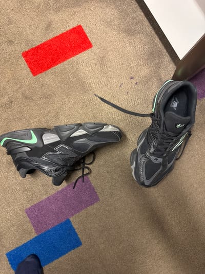 The image shows two New Balance sneakers, one model 9060 and the other model 1906r, on a carpet with rectangular colored sections of red, purple, and blue. A portion of a white-topped table is visible in the upper left corner, and part of a person's leg is visible in the lower right. There is no indication of the location of Katwijk, Netherlands.