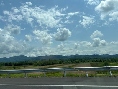 The image captures a landscape scene from Georgia, likely in or near the village of Chibati, within the municipality of Lanchkhuti. In the foreground, a paved road with a white line marking is visible, bordered by a metal guardrail. Beyond the guardrail, agricultural fields with patches of brown soil and green vegetation stretch towards rolling hills covered in dense green trees. In the background, a range of mountains forms the horizon, with the sky above filled with scattered white clouds against a bright blue backdrop. No people or specific actions are depicted in the image; it is a static view of the natural and agricultural environment.