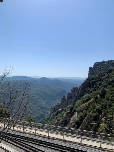 The image captures a panoramic view of a mountainous landscape, likely taken from a higher elevation, possibly near the city of Monistrol de Montserrat, Spain. In the foreground, there are several sets of railway tracks running horizontally across the frame, with a paved walkway and a metal railing situated above them. Bare tree branches extend into the upper left quadrant of the image, adding a natural, somewhat stark element to the scene.
The middle ground is dominated by a lush, green mountainside densely covered in trees. Prominent rocky outcrops and formations jut out from the terrain, creating a dramatic and rugged texture. The slopes of the mountains are layered, with a winding road or path visible in the valley below.
The background features a series of undulating mountain ranges that recede into the distance, becoming increasingly hazy and lighter in color due to atmospheric perspective. The sky is a clear, bright blue, indicating a sunny day. A small cross is visible on the summit of the furthest rocky peak on the right side of the image, suggesting a significant landmark or religious site. Electrical wires and infrastructure for the railway are also present, indicating the proximity of human activity and transportation.
