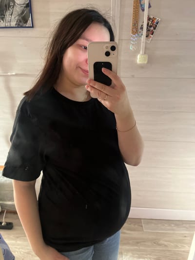 In an interior residential setting in Kozlovka, Russia, a female subject is depicted from the waist up, reflected in a mirror. She has dark brown hair and is wearing a black short-sleeved t-shirt and light blue jeans. Her right hand, adorned with a thin gold-colored ring on the ring finger and a thin gold-colored chain bracelet on the wrist, holds a light-colored iPhone vertically, obscuring the left side of her face. A dark accessory is affixed to the back of the phone. A faint silver-colored chain necklace is visible around her neck. The subject exhibits a slight smile and a prominent abdominal bulge, consistent with pregnancy. The background, visible in the mirror reflection, consists of a light-colored wall with horizontal paneling. A framed artwork with a dark blue border is visible on the upper left wall. On the upper right wall, a decorative wooden item with hanging elements and several small photographs are attached. The floor is covered with light wood-patterned material, and a white baseboard runs along the wall.