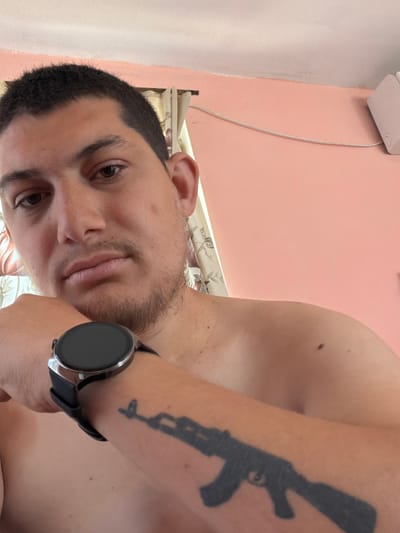 The media file is a close-up, rotated selfie of a young man. He is looking directly at the camera with a neutral expression. He has short, dark hair and a light beard. On his left arm, a dark tattoo of a rifle is visible. He is wearing a smartwatch on his left wrist. The background is a pastel pink wall with a white wire and an outlet visible. A patterned fabric, possibly bedding or curtains, is also visible. The scene appears to be indoors, with soft lighting. There is no visible text in the image. The image was taken in Biser, Bulgaria.