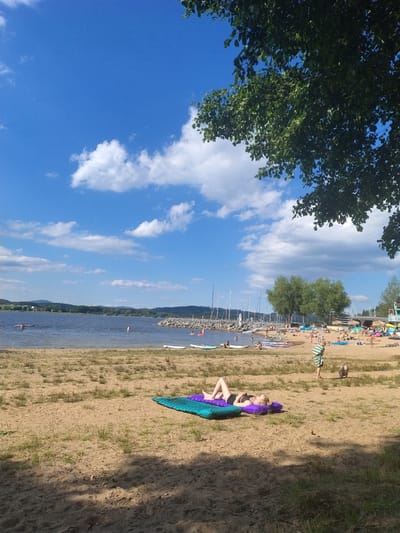 The image depicts a crowded beach scene on a sunny day, located at the Radslav Reservoir in Radslav, Czechia. The foreground is dominated by a sandy beach with sparse patches of grass. Two individuals are visible in the immediate foreground, with one woman lying on a purple and blue inflatable mat, sunbathing. Another person, a child, is also present in the sand. In the middle ground, the beach extends towards the water, lined with numerous people, umbrellas, beach chairs, and various watercraft, including paddleboards and kayaks. The water of the reservoir is calm, with several people swimming or wading. Across the water, a treed shoreline is visible, leading to rolling hills in the distance. The sky is a vibrant blue, dotted with white, fluffy clouds, and partially obscured by the dark green foliage of a large tree in the upper right corner of the frame. Sailboats are docked along a rocky breakwater in the mid-ground on the left side of the reservoir.