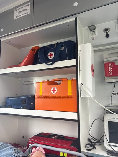 The image shows the interior of an ambulance, with medical supplies neatly organized on shelves. On the top shelf, there is a blue medical bag with a red cross emblem, and to its left, an orange tubular object. A white sticker on the cabinet above reads "Воротник Шанца Небулайзер Шины мягкие" which translates to "Shants collar, Nebulizer, Soft splints."
The middle shelf contains an orange medical case with a red cross, a yellow stripe, and the text "МЕДПЛАНТ" printed on it. To the left of this case is a blue bag with metal stud details. A white sticker on the shelf above reads "Комплект для дефибриллятора" which translates to "Defibrillator kit."
The bottom shelf holds a red bag with black accents, and to the left, a person's legs are visible, wearing blue jeans with a speckled pattern.
On the right side of the ambulance interior, a white device with a red cross emblem is mounted on the wall. Below it, a red package labeled "ACCUVAC Rescue" is visible. Further down, a monitor screen with the brand name "AXION" is present, connected by cables. Electrical outlets and other medical equipment are also visible. The image appears to have been taken in Pesochny, Russia, as indicated by the context provided.