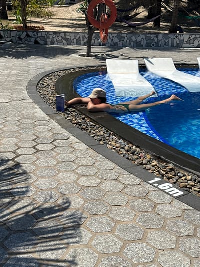 A person wearing a bathing suit and hat relaxes poolside, legs in the water. They are reading a blue book. The pool edge is marked "1.6m." The pool is lined with blue and white tiles. The poolside area is paved with gray hexagonal stones. Tree shadows fall across the paving. A life preserver hangs nearby.