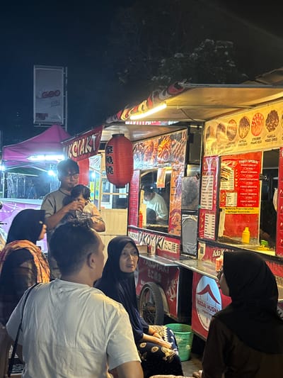 The image depicts a lively night scene at a street food stall, likely in Tanggul, Indonesia. The stall, labeled "TAKOKYAKI," is brightly lit and displays a variety of food items and prices on its signage. Several people are gathered around the stall, some appearing to be customers and others potentially staff.
In the foreground, a man wearing a white t-shirt is seen from behind, facing the stall. To his right, a woman in a black hijab is looking towards the stall with a thoughtful expression. To the left of the man, a woman with a patterned scarf and hijab is partially visible. A man holding a young child stands near the stall's counter. The child, held by the man, appears to be looking down.
The stall itself has a colorful awning with red, white, and black patterns. A large red lantern hangs prominently from the stall's roof. The signage features various food options like "CAPPUCCINO," "NUTELLA," "STROBERI," and "VANILLA MESES OREO," along with prices in Indonesian Rupiah. There are also menus displayed with item names and prices, and posters indicating "PAKET SPESIAL" (Special Package). One poster lists "PAKET COKLAT," "PAKET STROBERI," and "PAKET TIRAMISU."
In the background, another stall with a pink canopy is visible. A tall sign with "geo mild" branding is mounted on a pole. The overall environment suggests a bustling street market or food court at night, with the presence of trees and a dark sky overhead. A green plastic basket is placed on the ground near the seated woman. The details indicate a typical Indonesian street food setting.