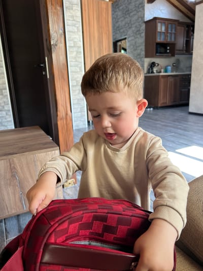 A young, fair-skinned, blond-haired child is looking down, reaching into a red and maroon checkered backpack. The child is wearing a tan-colored, long-sleeved shirt. The background includes brown wooden cabinets, a gray brick wall, and a wooden countertop. Based on the interior design elements such as the cabinets and brick wall, this is a domestic environment. While the image does not explicitly show outdoor landmarks, and considering the provided location, the image appears to be taken indoors in a residential area of Gradezhnitsa, Bulgaria.