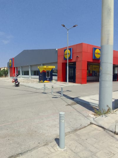 This media file depicts a bright, sunny day in Aspropyrgos, Greece, outside a Lidl supermarket. The building has a modern architectural style with a gray facade and bright red accents around the entrance. The iconic yellow and red Lidl logo is prominently displayed on the exterior.

In front of the store, an ATM machine is visible, colored yellow, blue, and red, with Greek text indicating "ATM ΧΩΡΙΣ ΧΡΕΩΣΗ" (ATM Without Charge). A parked motorcycle, a common mode of transportation in Greece, is positioned to the left of the entrance, suggesting recent activity or a customer's arrival. A tall concrete pole stands in the foreground, casting a shadow on the paved area. The ground is a mix of asphalt and paved walkways, with some greenery growing along the edges, indicating a typical urban or commercial environment.

The sky is a clear, vibrant blue, and the sunlight suggests it is daytime, likely late morning or early afternoon given the strong shadows. The overall scene conveys a sense of everyday commerce in a Mediterranean setting. There are no people clearly visible in the image, but their presence is implied by the parked motorcycle and the operational ATM. The scene appears tranquil and uncrowded.