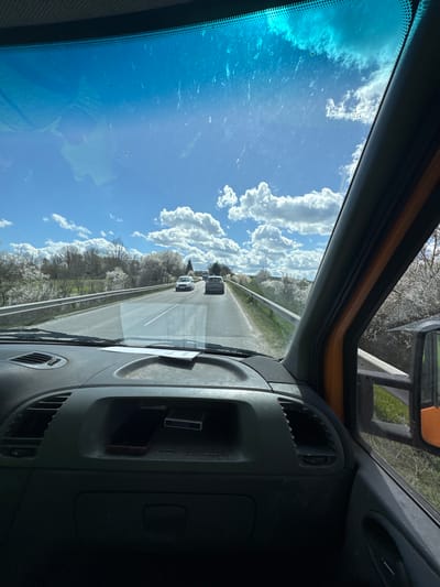 The image depicts a view from inside a vehicle, likely a truck, driving on a highway. Outside the windshield, there's a clear blue sky with puffy white clouds. Trees with white blossoms line the sides of the road. Two cars are visible ahead on the highway. The dashboard of the vehicle is visible, including air vents and a small compartment. The photograph doesn't provide enough information to determine the exact location within Venets, Bulgaria.