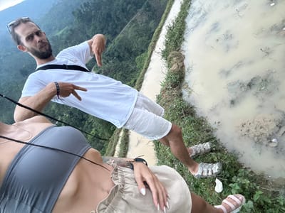An eye-level outdoor shot, rotated 90 degrees clockwise, captures two individuals amidst muddy fields and verdant hills in Nậm Dịch Commune, Vietnam.
In the right foreground, a man, shown from the waist up, poses for the camera. He wears a white t-shirt, light-colored shorts, and grey sandals. He has a beard and sunglasses resting on his head. With both hands, he forms a "peace" or "V" sign. A black strap, likely from a bag, crosses his chest. His expression is neutral.
On the left side of the frame, parts of a woman are visible, from her feet to her torso. She wears pink sandals and light-colored shorts. Her hand, adorned with long white nails, rests near her hip, revealing a bracelet and a tattoo on her arm. She is wearing a grey bra-top, and a thin necklace is draped over her chest.
The immediate surroundings consist of muddy, waterlogged terrain, resembling rice paddies or flooded agricultural fields, bordered by green grass and dirt paths. In the distant background, lush green mountains rise, appearing somewhat hazy or misty, suggesting an overcast day. The scene depicts a casual outdoor activity in a rural, mountainous landscape. No discernible text is visible in the image.