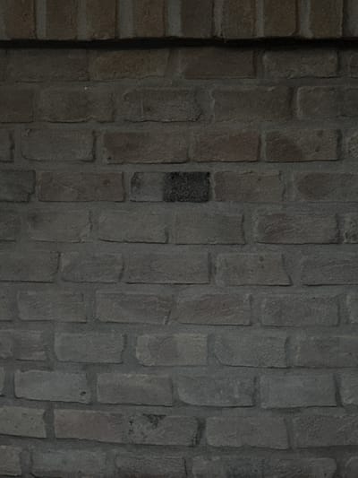 This media file is a close-up, textured image of a dark, weathered brick wall. The bricks are rectangular and laid in a staggered pattern with visible mortar lines. The color palette is predominantly grays and dark browns, suggesting an aged material and potentially low light conditions. There are no people or discernible objects in the frame, only the wall itself. The surface of the bricks exhibits variations in texture and color, with some appearing smoother and lighter than others, indicating wear and tear over time. One brick towards the center-left of the image is notably darker than its surrounding counterparts, possibly due to discoloration or a different material composition. The lighting is subdued, creating subtle shadows that highlight the three-dimensional quality of the brickwork and mortar. There is no visible text in the image. The setting appears to be an outdoor or indoor wall, given the rustic nature of the brick. Without further context, specific details about the time of day, weather, or emotional tone are difficult to ascertain, though the overall impression is one of age and perhaps a somber or neutral atmosphere. The location context of Sant'Angelo di Piove di Sacco, Italy, implies that this brick wall is part of the architectural landscape of this Italian town.