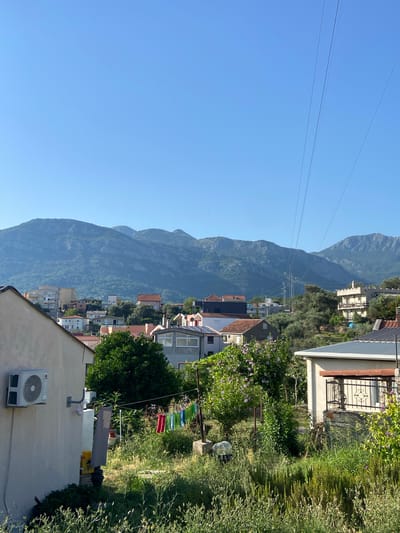 This image captures a view of Burtaiši, Montenegro, characterized by a cluster of residential buildings nestled against a backdrop of lush green hills and distant mountains under a clear blue sky. In the foreground, a light-colored house with an air conditioning unit mounted on its exterior wall is visible on the left. Adjacent to it, laundry is hanging on a clothesline, with several brightly colored garments, including red and green towels, and blue and white clothing.

The midground reveals a densely packed residential area with various houses, many featuring red-tiled roofs. These homes are interspersed with trees and vegetation, creating a vibrant, organic feel. Some houses have visible balconies or windows, suggesting the presence of inhabitants. The architecture appears to be a mix of traditional and more modern styles.

In the background, imposing mountains dominate the horizon, their slopes covered in dense greenery. The sky is a uniform, cloudless blue, indicating fair weather. Overhead, power lines stretch across the sky, a utilitarian element contrasting with the natural beauty of the landscape. The overall impression is one of a semi-rural or suburban setting, where residential life coexists with a striking natural environment.