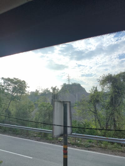 The image depicts a roadside scene in Bilaspur, India, on a cloudy day. In the foreground, a gray asphalt road with a white dividing line stretches across the bottom of the frame. A silver metal guardrail runs parallel to the road, separating it from a rocky embankment covered in greenery. Beyond the embankment, lush trees and vegetation dominate the landscape, with a prominent transmission tower visible on a distant hilltop. The sky is partly cloudy, with patches of blue visible through the white clouds. A dark, tinted visor or dashboard is visible at the top of the frame, suggesting the photo was taken from inside a vehicle. A tall, slender pole with a rectangular sign is positioned near the guardrail.