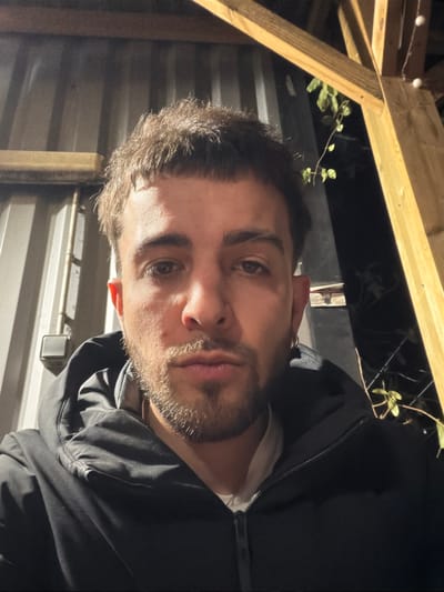 A man with short, dark hair and a trimmed beard looks into the camera. He is wearing a black hoodie and a white shirt. The background shows a wooden structure, possibly a pergola, with string lights and some greenery. A corrugated metal wall is also visible. The image is rotated, giving a tilted perspective.