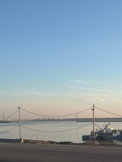 This is a vertical, eye-level, full shot image taken from a road, looking out over a body of water at dusk. The sky is a clear, pale blue that transitions to a pale orange hue near the horizon. A distant bridge spans the water, with industrial structures visible on the far bank. Two poles with wires are in the foreground, one on the left and one on the right. In the water, to the right of the image, are several boats docked together, including a white tugboat with a cabin and a blue hull. The water is calm and reflects the sky. The road in the foreground is dark asphalt. The overall scene suggests a port or industrial area.