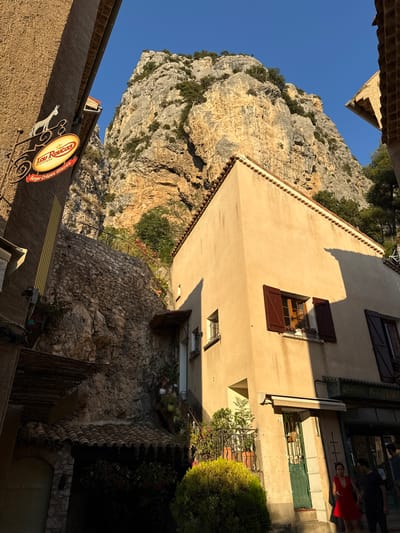 The image depicts a narrow street in Moustiers-Sainte-Marie, France, with a building situated against a dramatic rocky cliff face. The sun is low in the sky, casting long shadows and illuminating the stucco facade of the building with a warm, golden light.
On the left side of the street, a sign for "Lou Roucas" hangs from a metal bracket, featuring an oval logo with a star and the text "MOUSTIERS" and "Burger Crêpes Glaces." The sign is adorned with a silhouette of a donkey. Above this sign, part of another building's textured wall is visible, with a pitched roofline and a gutter.
The building in the foreground is a two-story structure with a cream-colored stucco exterior. It has multiple windows, some with dark brown shutters, and a tiled roof. A green door with a metal grate is visible on the ground floor, along with a small awning. Several potted plants and shrubs, including a dense green bush, are placed outside the building.
The backdrop is dominated by a sheer, rugged rock cliff, characteristic of the Verdon Gorge region. The cliff face is rough, with visible layers of rock and sparse vegetation growing from crevices. The sky above is a clear, bright blue.
In the lower right portion of the image, two individuals are walking on the sidewalk. A woman in a red dress is in the foreground, and a man in a dark t-shirt and pants is beside her. Further back, another person can be seen entering a shop. The shopfront has a green awning and large glass windows.
The overall impression is of a picturesque village nestled within a dramatic natural landscape, bathed in the soft light of late afternoon or early morning.