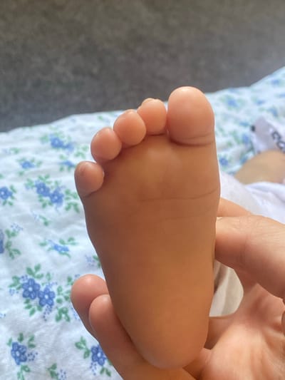 A close-up, overhead shot shows a human hand gently holding a newborn baby's left foot. The baby's foot is pink and plump with visible creases on the sole and five toes. The background features a white fabric with a blue floral pattern, likely a blanket or bedding, and a gray, textured surface, possibly a bedspread or a wall. The lighting suggests natural daylight. The scene is devoid of any context related to Arbagish, Uzbekistan.