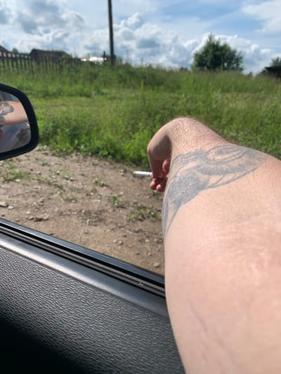 A first-person perspective shot shows a person's arm with a tattoo on it, resting on the open car door. A cigarette is held between the person's fingers. The scene appears to be outdoors, with a grassy field and a wooden fence visible in the background, suggesting a rural or suburban setting possibly in Barsai, Russia. The sky is partly cloudy. The car interior is visible in the foreground.