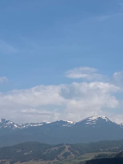The image shows a view of snow-capped mountains against a blue sky with white clouds. The mountains are partially covered in greenery, with some patches of snow visible on the peaks and slopes. The sky is mostly clear with some scattered clouds. The location is likely Kraishte, Bulgaria, given the request, but this cannot be verified solely from the image.